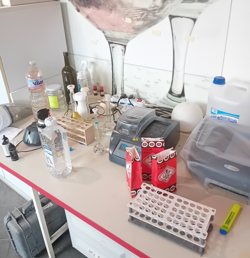 Banco di laboratorio con fotometro, provette in rack bianchi, matracci, sacchetti rossi e tanica di acqua demineralizzata